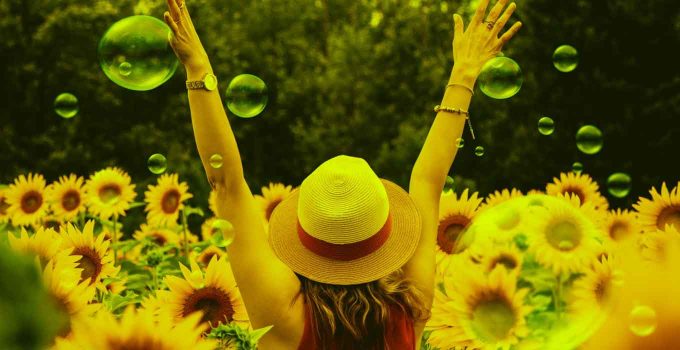 A woman with a hat raising her hands among sunflowers.